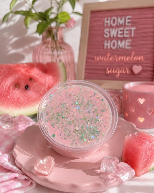 A wax melt in a clear round container sits on a pink plate, surrounded by watermelon slices, pink accessories, and a letter board that reads 'HOME SWEET HOME watermelon sugar'.