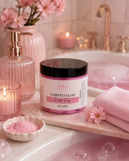 A jar of PWS Whipped Soap in 'Whipped Fantasy' is displayed on a marble bath tray next to a pink towel, bath salts, and a pink ribbed soap dispenser in a bubble-filled bathroom setting.
