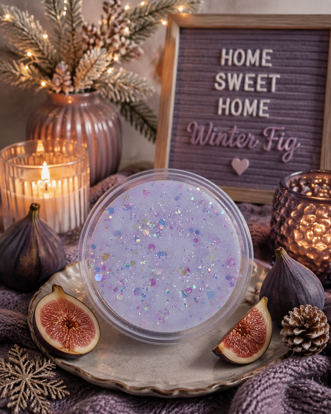 A round clear-lidded jar filled with pale lavender wax containing iridescent glitter, styled on a tray with fig halves, candles, a letter board reading 'HOME SWEET HOME Winter Fig,' and warm holiday decor.