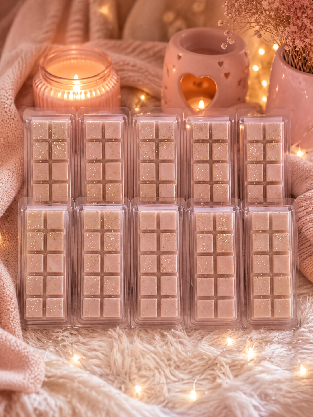 Ten wax melts in clear clamshell packaging, arranged in two rows of six on a soft white faux fur surface. The scene is styled with warm ambient lighting, including a lit candle, a pink heart-shaped holder, dried flowers, and string lights.