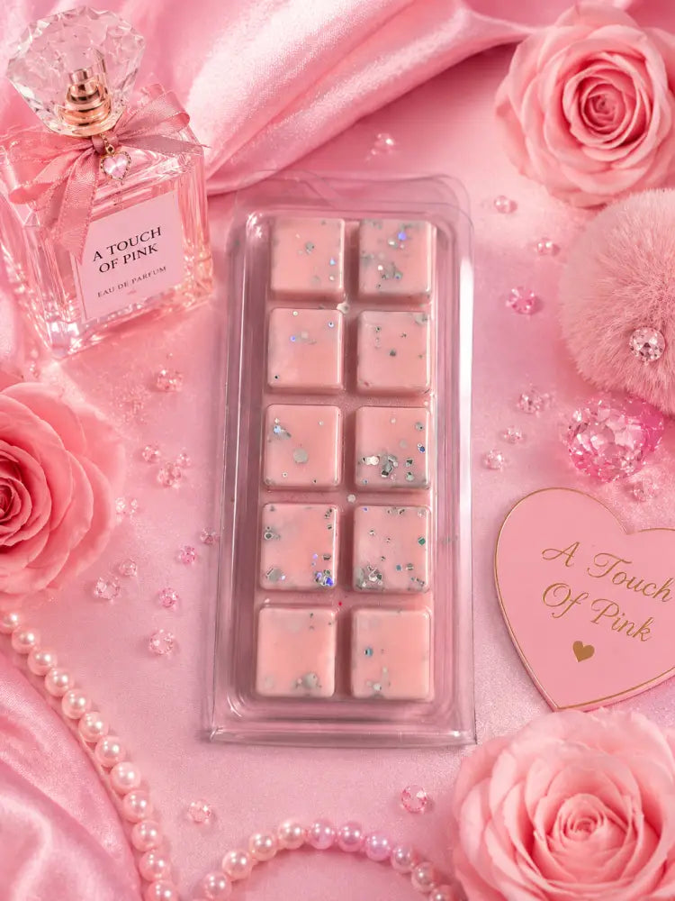 Pink wax melts in a clear plastic tray, styled on pink satin with pink roses, pearls, a perfume bottle labeled 'A Touch of Pink,' and a heart-shaped card reading 'A Touch of Pink.'.