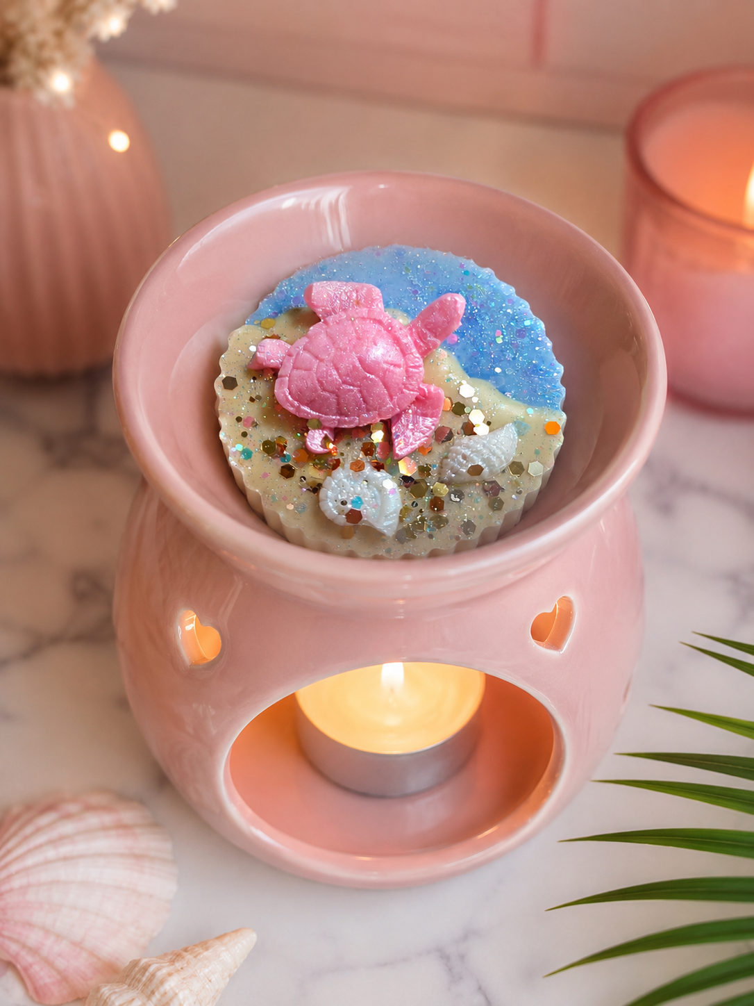 A pink ceramic wax warmer with a tealight candle inside, holding a round decorative wax melt topped with a pink sea turtle figure, blue glittery sand, and small shells. The scene is styled on a marble surface with shells, green leaves, and another blurred candle in the background.
